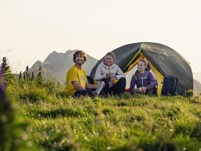 Drei Personen sitzen vor dem aufgebauten Zelt und betrachten das Bergpanorama.