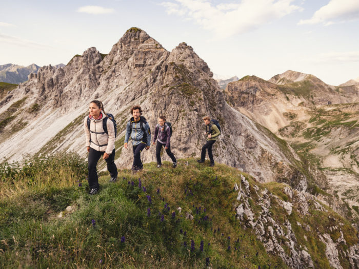 Vier Personen wandern auf einem Berggrat einen Weg entlang. Im Hintergrund sieht man eine Berglandschaft.